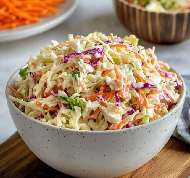 Close-up-shot-of-a-white-decorative-bowl-filled-with-crunchy-creamy-coleslaw-made-of-shredded-green-and-purple-cabbage-crisp-carrots-and-fresh-herb-garnishes-on-a-wooden-table-top-cookout-side-dishes 