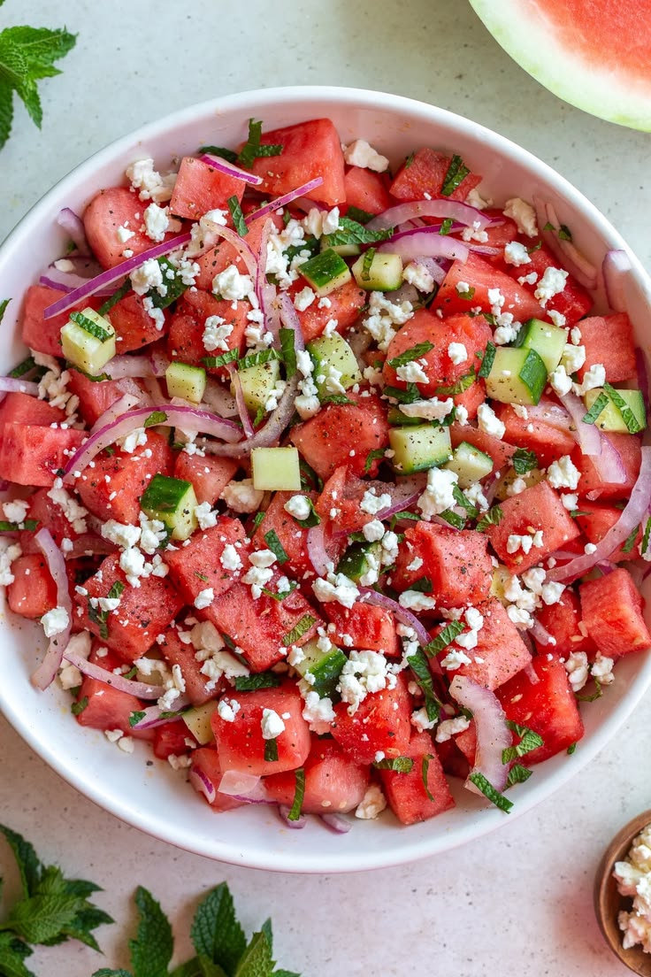 Bright-high-angle-view-of-a-white-bowl-filled-with-watermelon-feta-salad-featuring-diced-red-watermelon-cubes-crumbled-white-feta-cheese-chopped-green-mint-leaves-cucumber-pieces-and-sliced-red-onion 